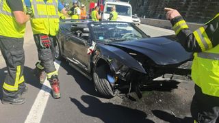 Ein beschädigtes schwarzes Sportwagen mit zertrümmerter Front steht am Straßenrand. Rettungskräfte in gelben Westen stehen darum herum.