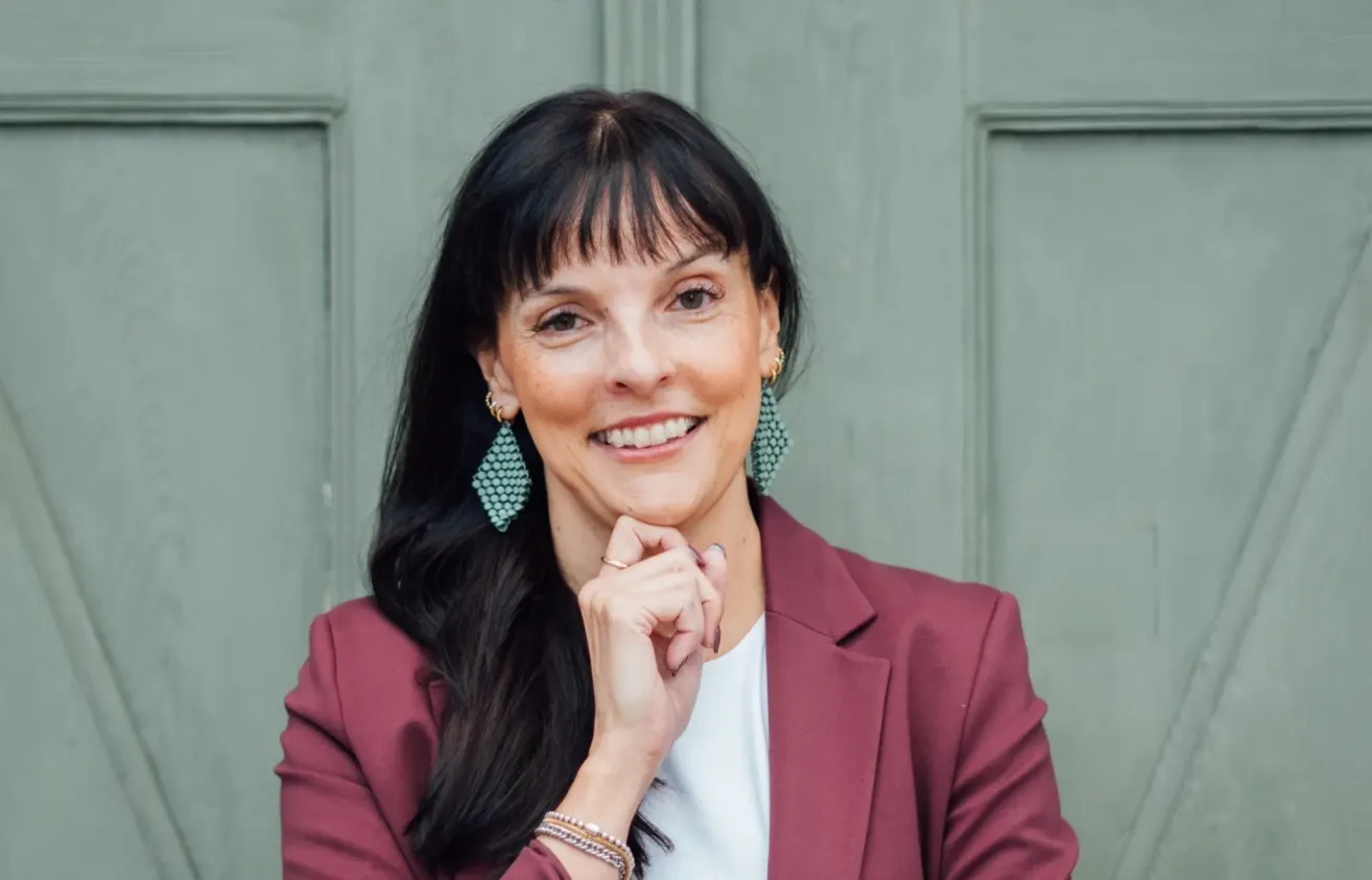 Eine Frau mit langen schwarzen Haaren, die einen kastanienbraunen Blazer und ein weißes Hemd trägt, lächelt und posiert für ein Foto.