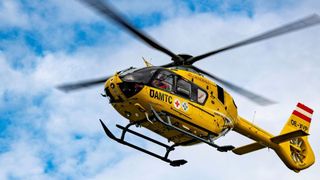 Ein gelber Hubschrauber mit DAMTC und rotem Kreuz fliegt am Himmel mit wolkigem Hintergrund.