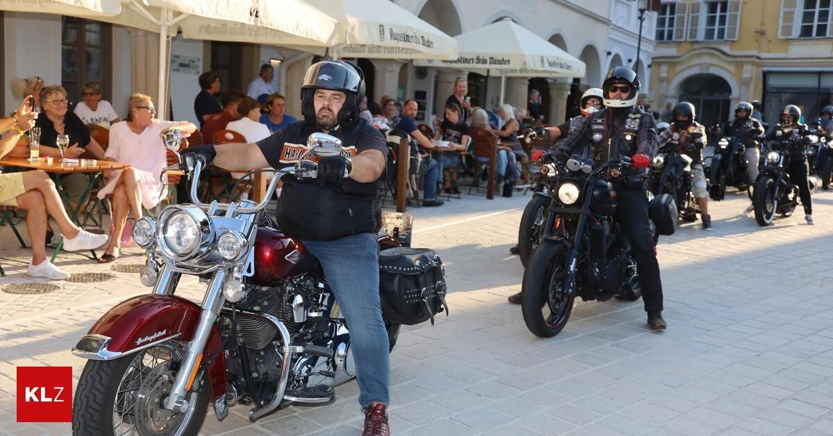 Eine Gruppe von Motorradfahrern mit Helmen und Lederjacken fährt auf einer gepflasterten Straße mit einem Café im Hintergrund.