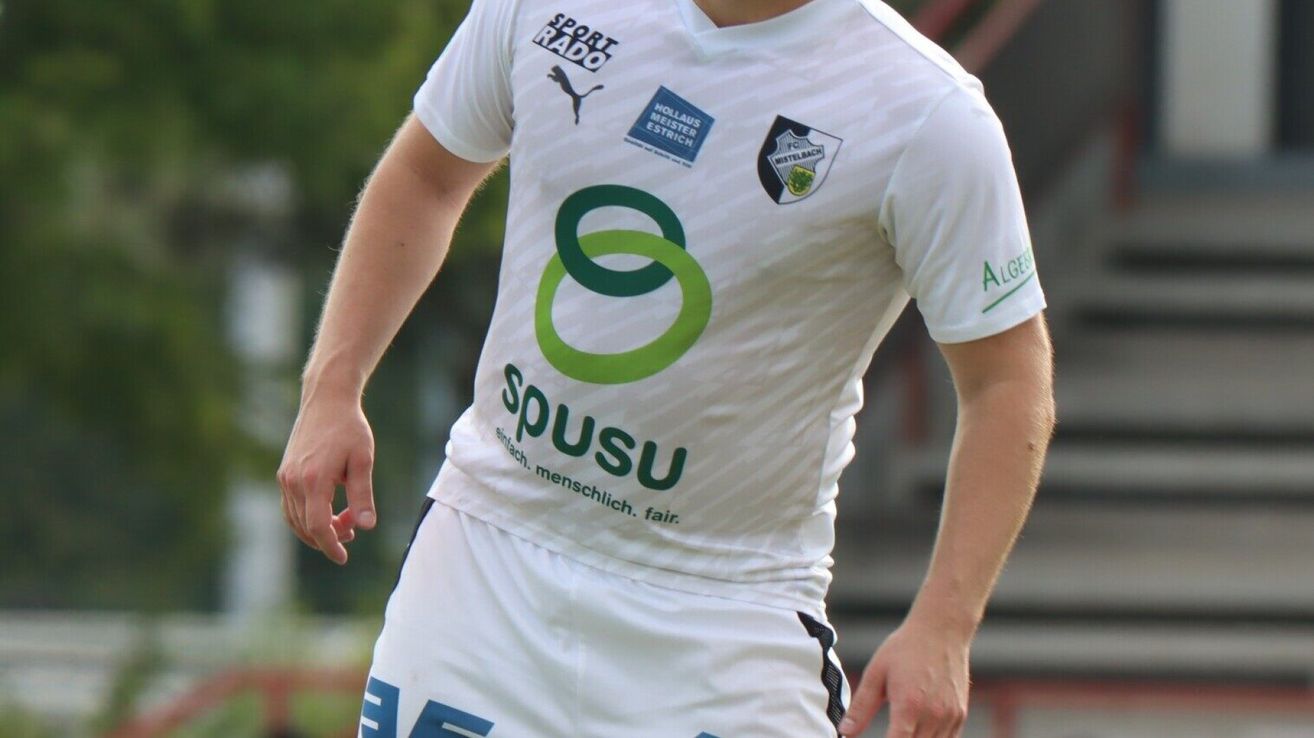 A soccer player wearing a white jersey with green and blue logos and text. The jersey has a sponsor logo on the front and a club logo on the right side.