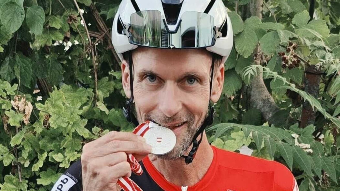 Ein Mann mit einem weißen Helm und einer silbernen Medaille um den Hals, lächelt und hält die Medaille in den Händen.