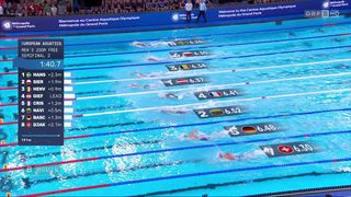 Schwimmer konkurrieren in einem Becken im Centre Aquatique Olympique in Paris, mit Zahlen und Flaggen, die ihre Positionen anzeigen.