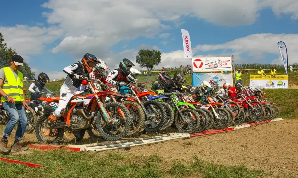 Eine Gruppe von Motocross-Fahrern in Helmen und Rennanzügen steht auf einer Schlammpiste in Reihe, mit Bannern und Zuschauern im Hintergrund.