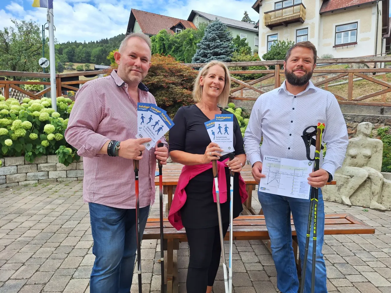 Drei Personen stehen vor einer Holzbank, jede hält einen Wanderplan und ein Paar Trekkingstöcke. Der Mann links trägt ein rosa Hemd, die Frau in der Mitte ein schwarzes Oberteil und der Mann rechts ein weißes Hemd. Hinter ihnen steht ein Haus mit Balkon und Zaun.