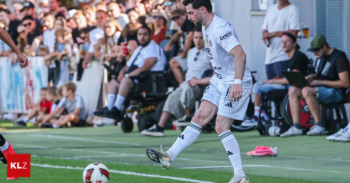 Ein Fußballspieler in einem weißen Trikot tritt einen Fußball auf einem Feld, während eine Menge zuschaut.