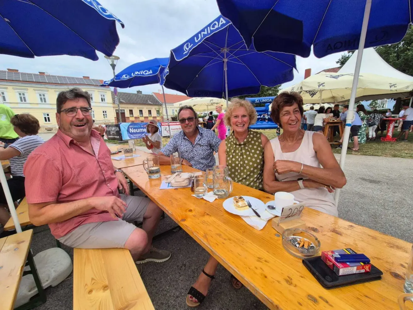 Vier Personen sitzen an einem Picknicktisch mit blauen Schirmen, lächeln und posieren für ein Foto. Der Tisch hat Gläser, Teller und ein Schild. Dahinter befinden sich Gebäude, ein Spielplatz und Menschen.