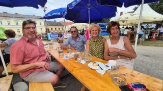 Vier Personen sitzen an einem Picknicktisch mit blauen Schirmen, lächeln und posieren für ein Foto. Der Tisch hat Gläser, Teller und ein Schild. Dahinter befinden sich Gebäude, ein Spielplatz und Menschen.