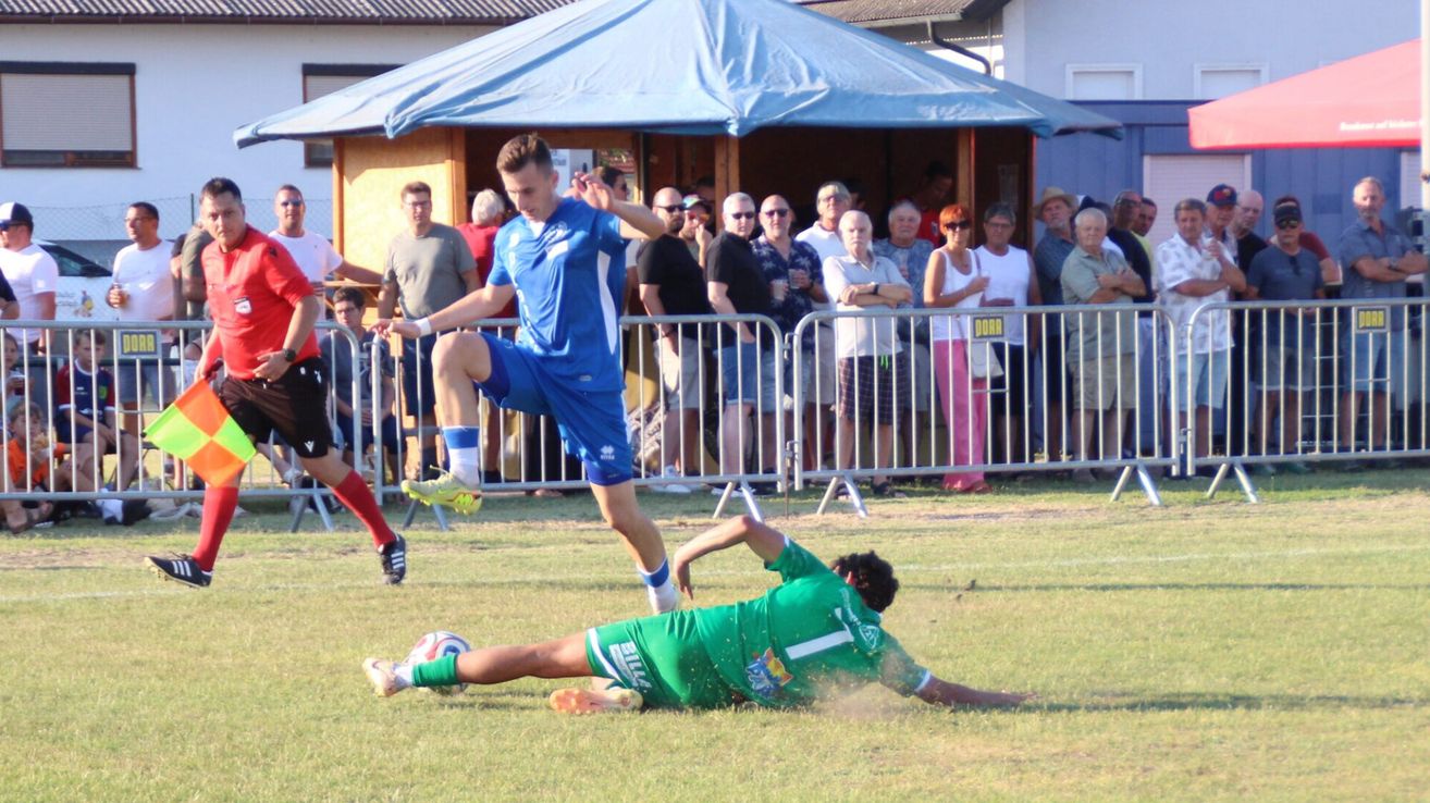 A soccer player in blue kicks the ball while another player in green attempts to block the shot. Spectators watch from behind a metal fence.