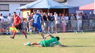 Ein Fußballspieler in Blau tritt den Ball, während ein anderer Spieler in Grün versucht, den Schuss zu blockieren. Zuschauer beobachten hinter einem Metallzaun.