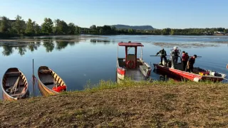 Einige Boote sind am Ufer eines Sees verankert, mit Menschen an Bord. Ein Boot hat ein rotes Dach.