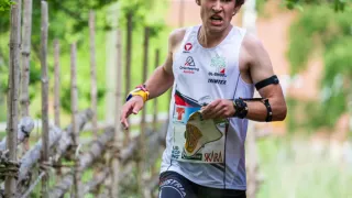 Ein Mann läuft einen Marathon und trägt ein weißes ärmelloses Shirt mit verschiedenen Logos und einem schwarzen Armband. Er hält ein Stück Papier in der rechten Hand.