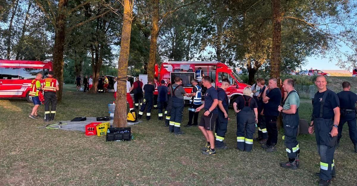 Eine Gruppe von Feuerwehrleuten in Uniformen versammelt sich um einen roten Lastwagen in einem Park, einige stehen in der Nähe eines Baumes und andere in der Nähe von Kisten.