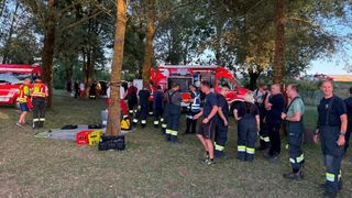 Eine Gruppe von Feuerwehrleuten in Uniformen versammelt sich um einen roten Lastwagen in einem Park, einige stehen in der Nähe eines Baumes und andere in der Nähe von Kisten.