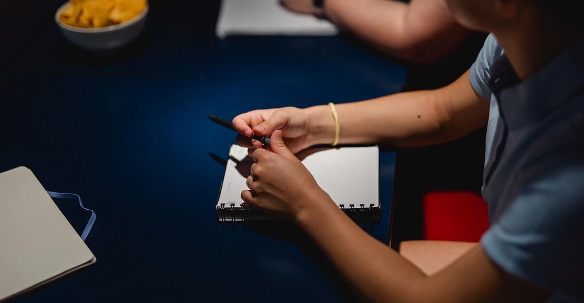 A person is writing on a notebook with a pen on a blue table. The person is wearing a bracelet.