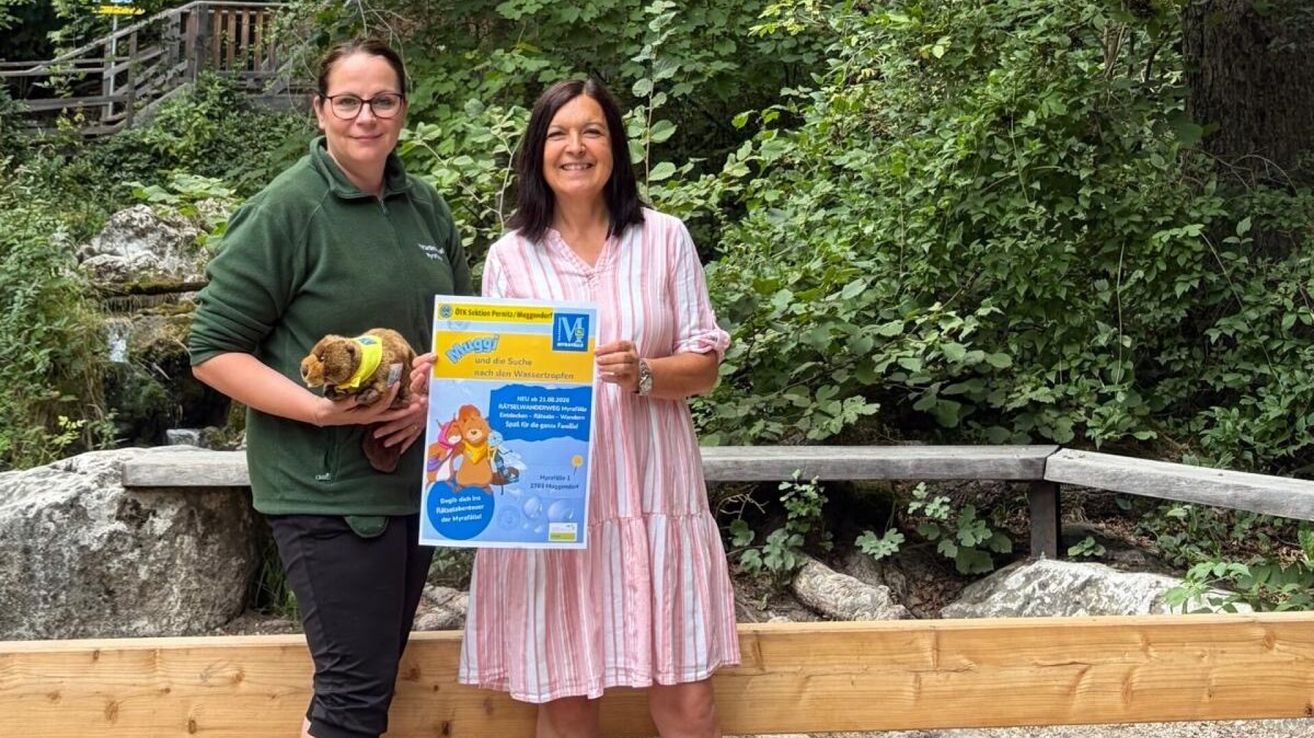 Zwei Frauen stehen in einem Wald, eine hält ein Kuscheltier und ein Plakat, die andere lächelt und hält das Plakat. Das Plakat zeigt einen Comic-Charakter und einige Texte. Hinter ihnen steht ein Holzzaun und Grünpflanzen.