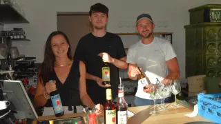 Drei Personen stehen hinter einer Bartheke, lächeln und halten Flaschen mit Alkohol in den Händen. Die Frau links hält eine Flasche Aparru, der Mann in der Mitte hält eine Flasche Aparru und der Mann rechts hält eine Flasche Monin.