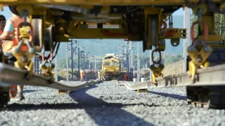 Ein Blick von unten auf einen gelben Zug auf einer Eisenbahnstrecke mit Arbeitern in orangefarbenen Uniformen in der Nähe.