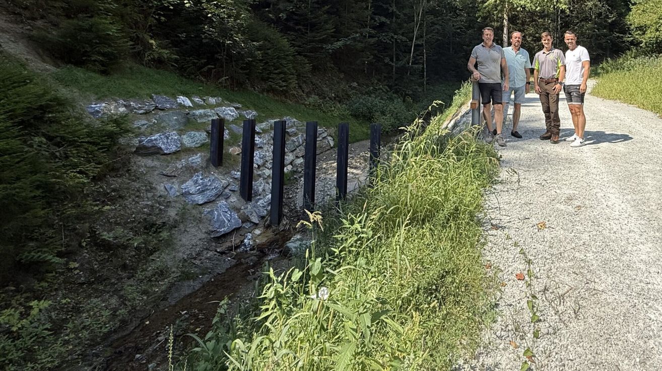 Zwei Männer stehen auf einem Schotterweg in der Nähe eines Waldes, mit einem kleinen Bach und Steinbarrieren auf der linken Seite.
