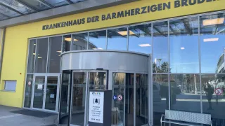 Der Eingang zu einem Krankenhaus namens Krankenhaus der Barmherzigen Brüder, mit einer Drehtür, einem Schild und einer Bank davor.