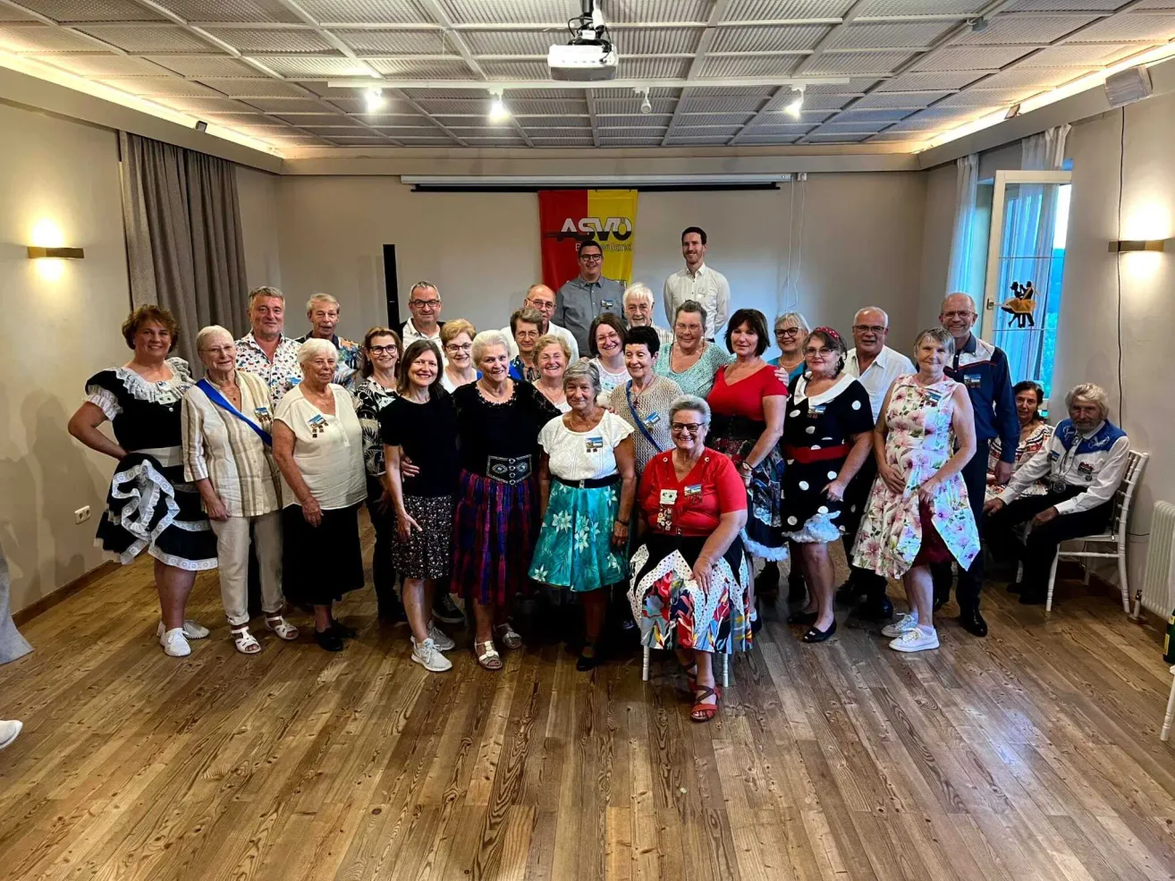 Eine Gruppe von Menschen, meist älterer, posiert für ein Foto in einem Raum mit Holzböden und einem Banner. Sie sind in verschiedenen Outfits gekleidet, einige tragen Brillen und Accessoires.