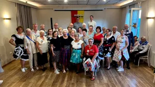 Eine Gruppe von Menschen, meist älterer, posiert für ein Foto in einem Raum mit Holzböden und einem Banner. Sie sind in verschiedenen Outfits gekleidet, einige tragen Brillen und Accessoires.