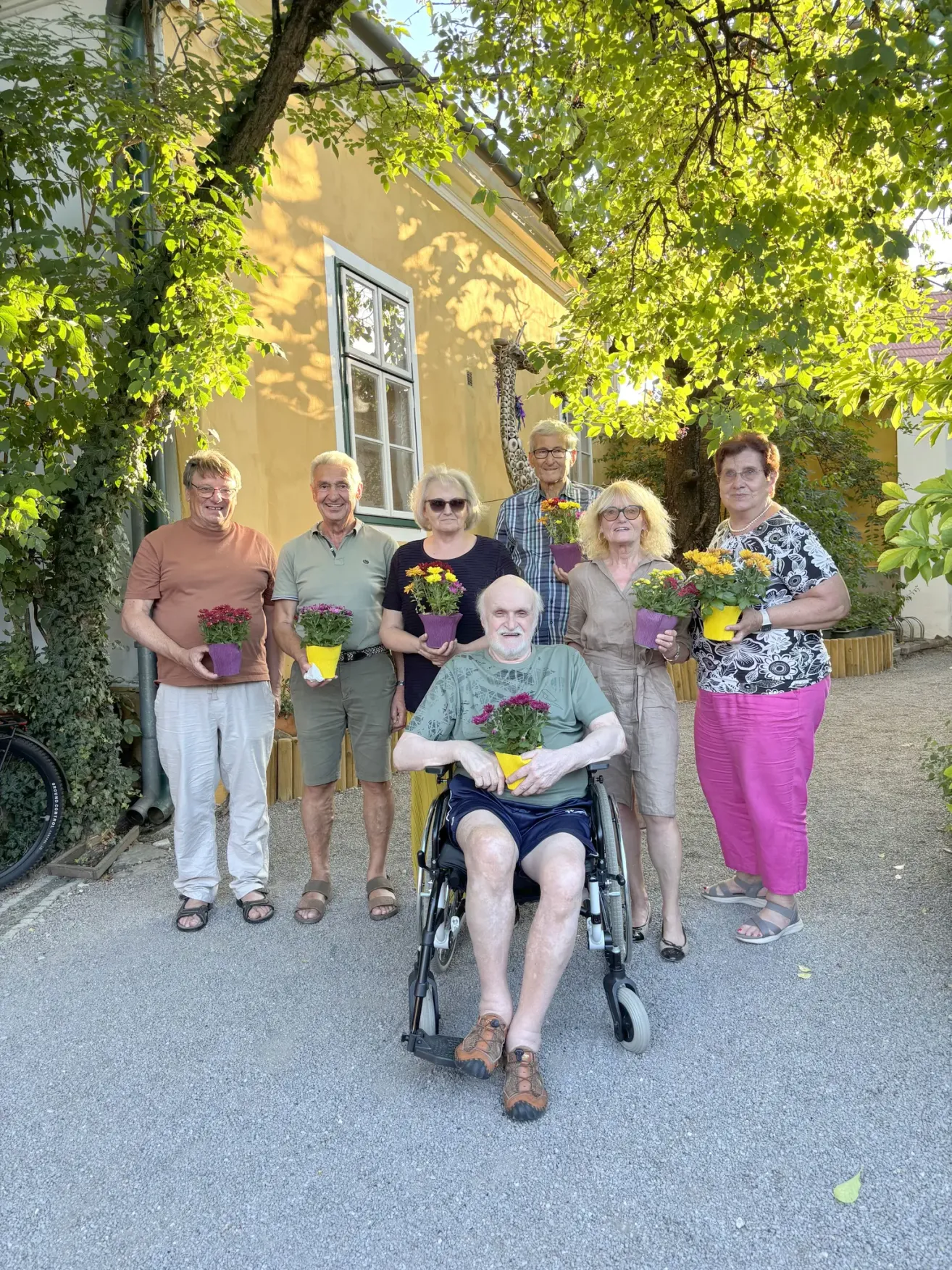 Eine Gruppe älterer Menschen steht vor einem gelben Haus, jeder hält eine Topfpflanze. Eine Person sitzt im Rollstuhl.