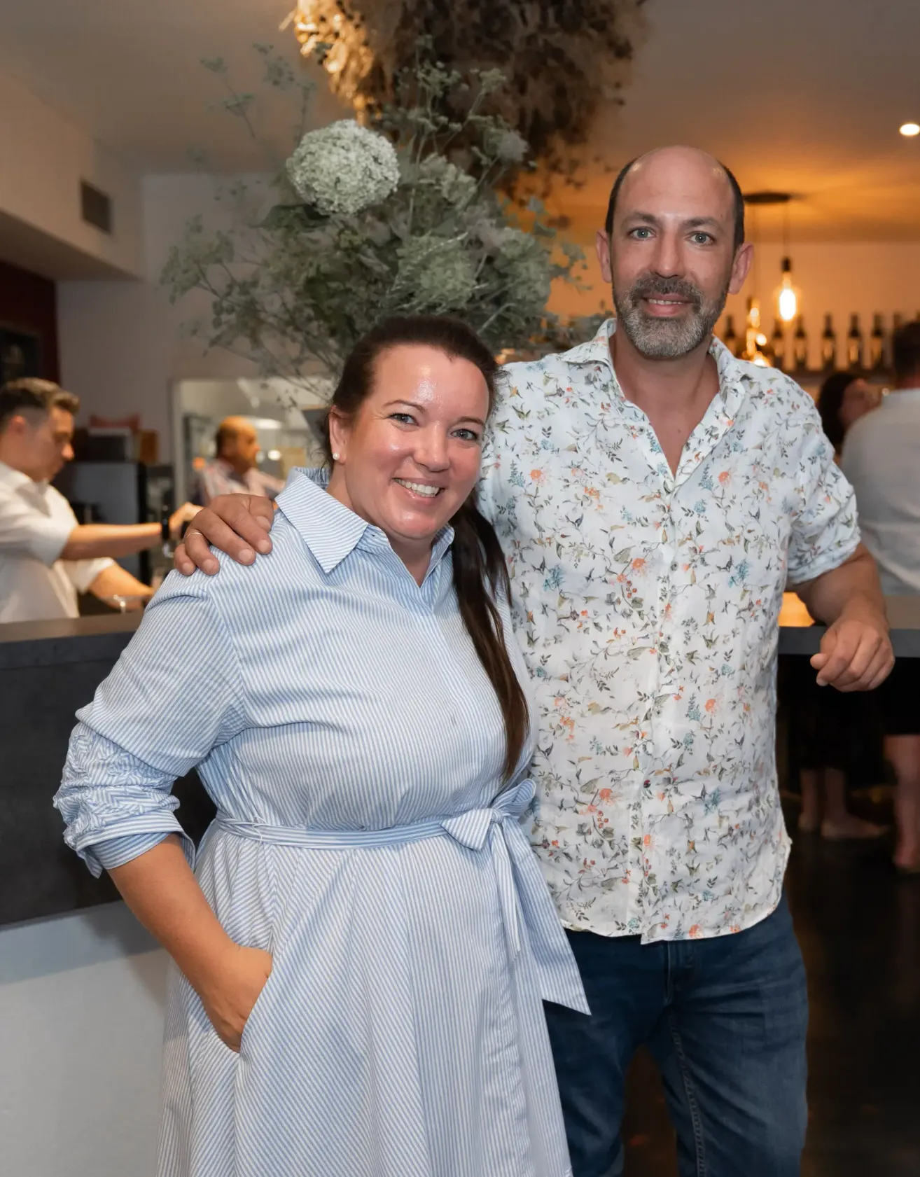 Ein Mann und eine Frau stehen lächelnd eng beieinander in einem Restaurant mit einem Blumenhemd und einem blauen Kleid. Hinter ihnen steht ein Mann an einer Theke.