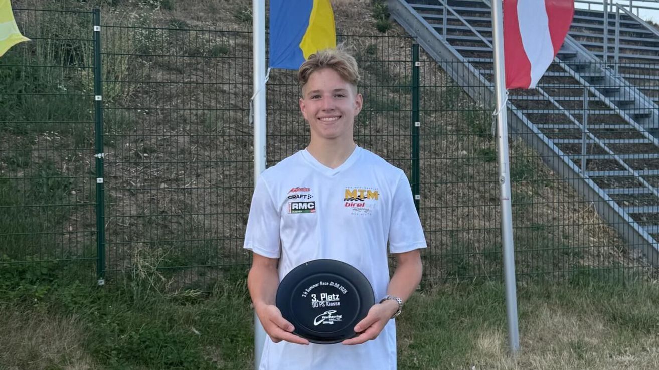 Ein junger Mann in einem weißen Shirt mit Logos steht vor einem Zaun und hält einen schwarzen Frisbee mit den Texten '3. Platz' und '90 PS Kasse'. Er lächelt und scheint sich bei einer Sportveranstaltung zu befinden.