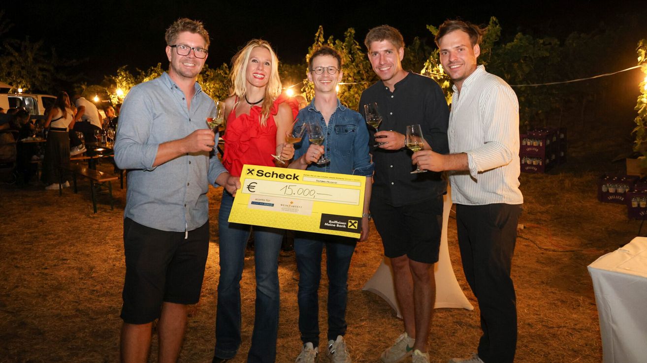 Fünf Personen stehen zusammen, lächeln und halten Weingläser. Eine Person hält einen großen gelben Scheck über 15.000 Euro. Sie sind draußen bei Nacht.