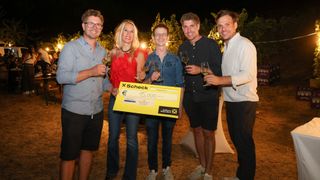 Fünf Personen stehen zusammen, lächeln und halten Weingläser. Eine Person hält einen großen gelben Scheck über 15.000 Euro. Sie sind draußen bei Nacht.