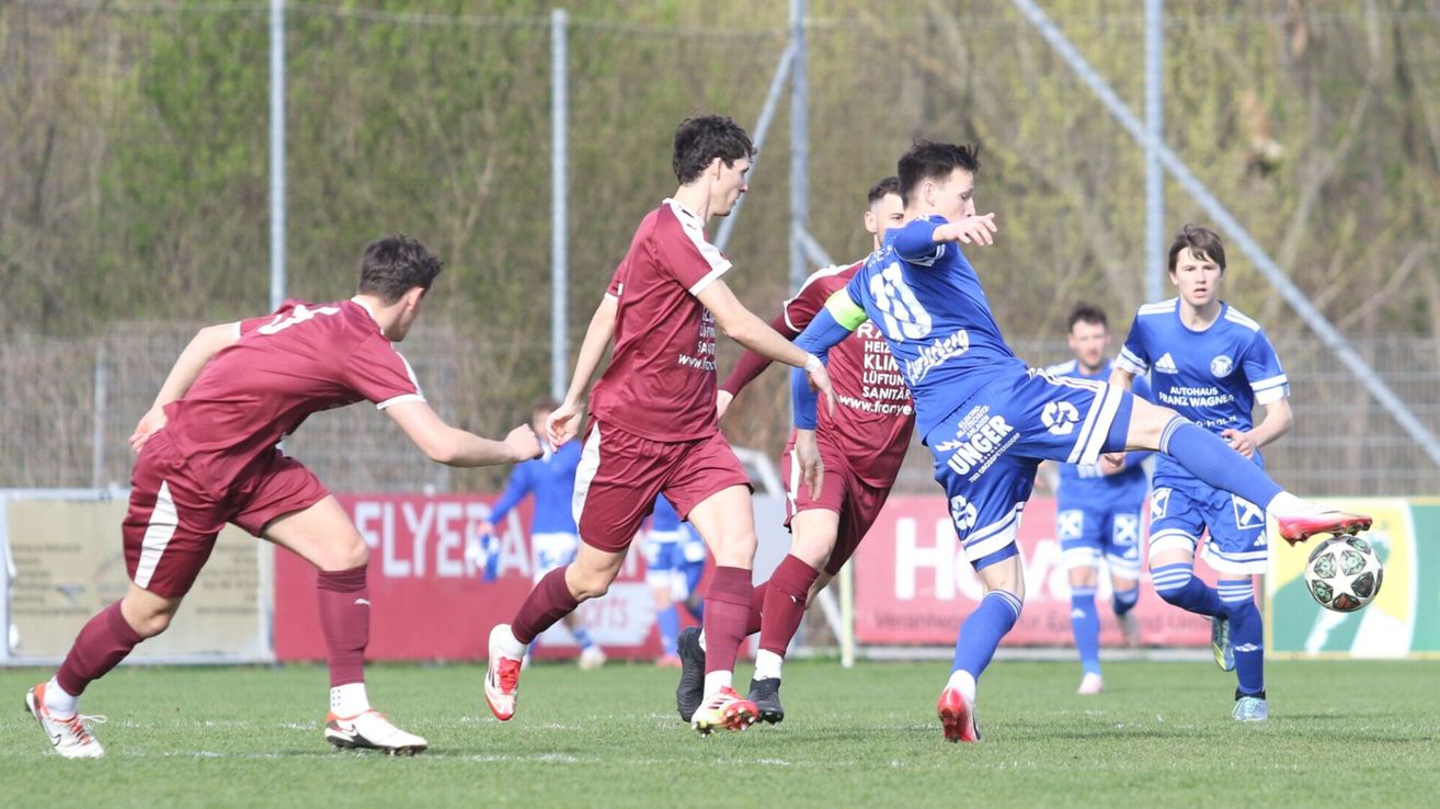 Ein Fußballspiel mit Spielern in burgunderroten und blauen Trikots, ein Spieler in Blau versucht, den Ball zu schießen, während ein anderer in Burgunderrot versucht, ihn zu blockieren.