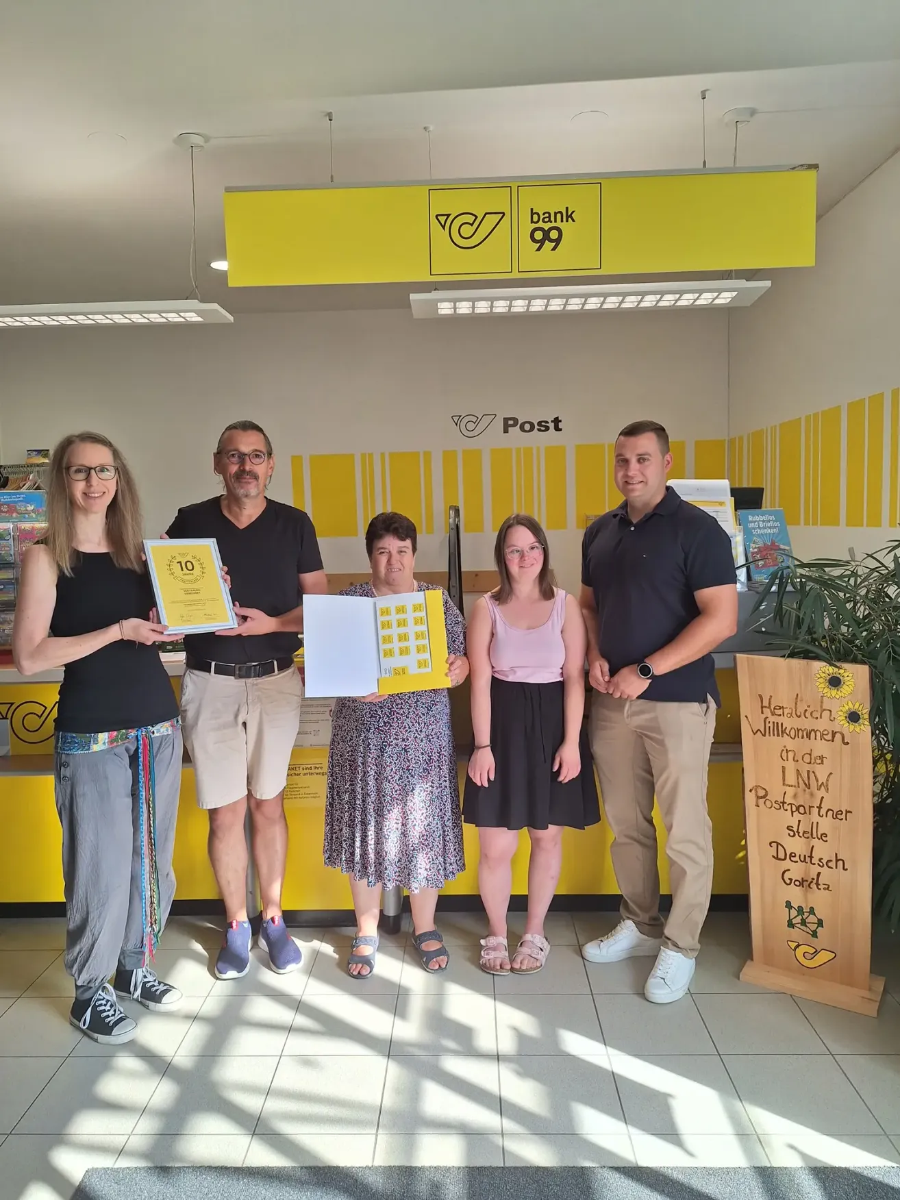 Five people stand in a room with yellow walls and a sign that says Post. They are holding certificates and smiling.