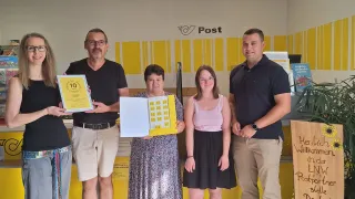 Fünf Personen stehen in einem Raum mit gelben Wänden und einem Schild, das Post sagt. Sie halten Urkunden und lächeln.
