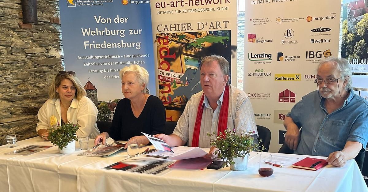 Drei Personen sitzen an einem Tisch mit Werbematerialien und einem Banner im Hintergrund. Der Mann hält ein Papier in der Hand und scheint zu sprechen.