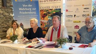 Drei Personen sitzen an einem Tisch mit Werbematerialien und einem Banner im Hintergrund. Der Mann hält ein Papier in der Hand und scheint zu sprechen.