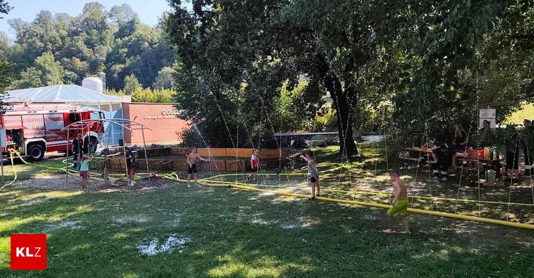 A group of people are playing in a water park with water sprinklers and a yellow hose on the grass. Trees and a building are in the background.