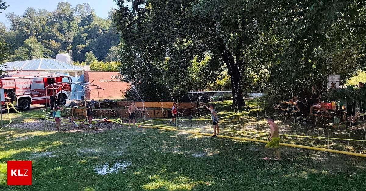 A group of people are playing in a water park with water sprinklers and a yellow hose on the grass. Trees and a building are in the background.