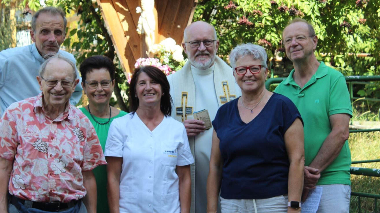 Eine Gruppe von Menschen, einschließlich eines Priesters, steht lächelnd für ein Foto in einem Garten.