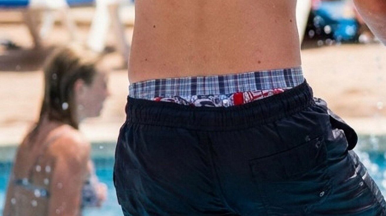 Eine Nahaufnahme des Rückens einer Person, die schwarze Badeshorts mit kariertem Bund trägt, in der Nähe eines Pools.