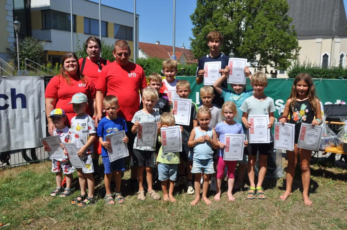 Eine Gruppe von Kindern und Erwachsenen steht auf einem Rasen und hält Urkunden in den Händen. Hinter ihnen ist ein grüner Banner teilweise sichtbar.