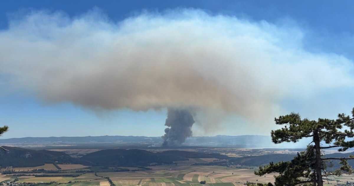 A large plume of smoke rises from a wildfire in a rural landscape, with hills and farmland visible in the background.