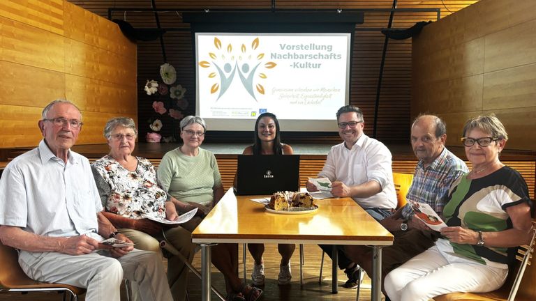 Eine Gruppe von Menschen sitzt um einen Tisch mit Laptop und Kuchen und lächelt. Ein Präsentationsbildschirm hinter ihnen zeigt 'Vorstellung Nachbarschafts-Kultur'.