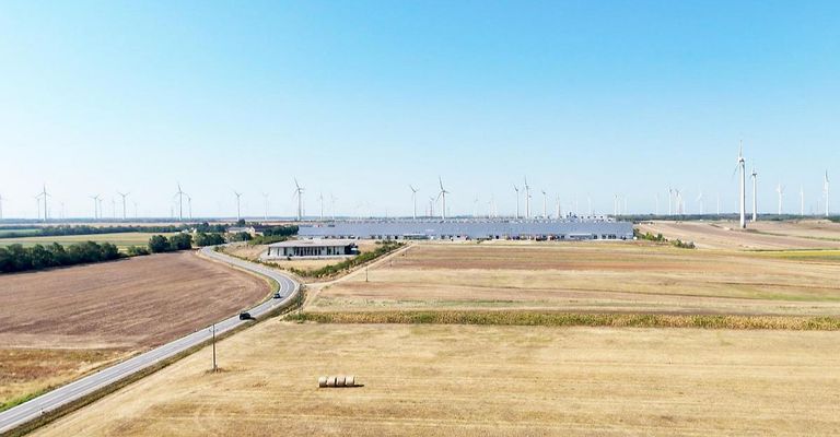 Eine Luftaufnahme einer großen Industrieanlage, umgeben von Windturbinen und einem weitläufigen, trockenen Feld unter einem klaren Himmel.