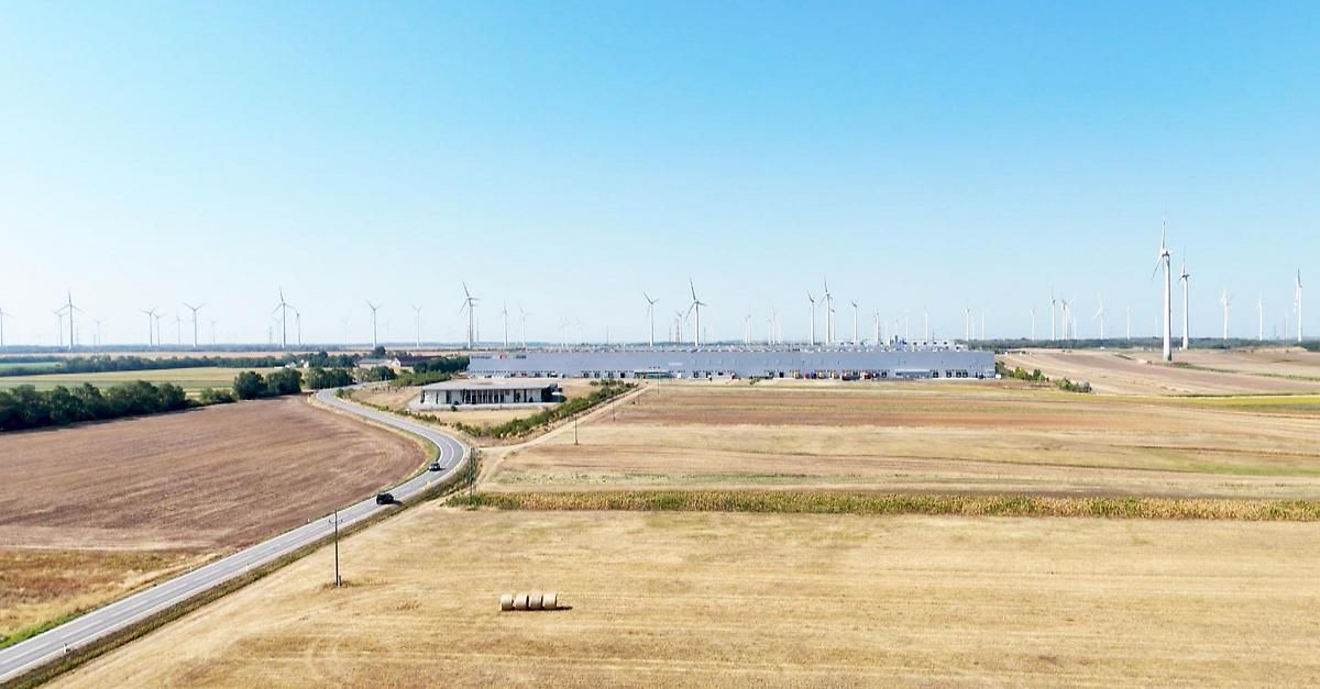 Eine Luftaufnahme einer großen Industrieanlage, umgeben von Windturbinen und einem weitläufigen, trockenen Feld unter einem klaren Himmel.