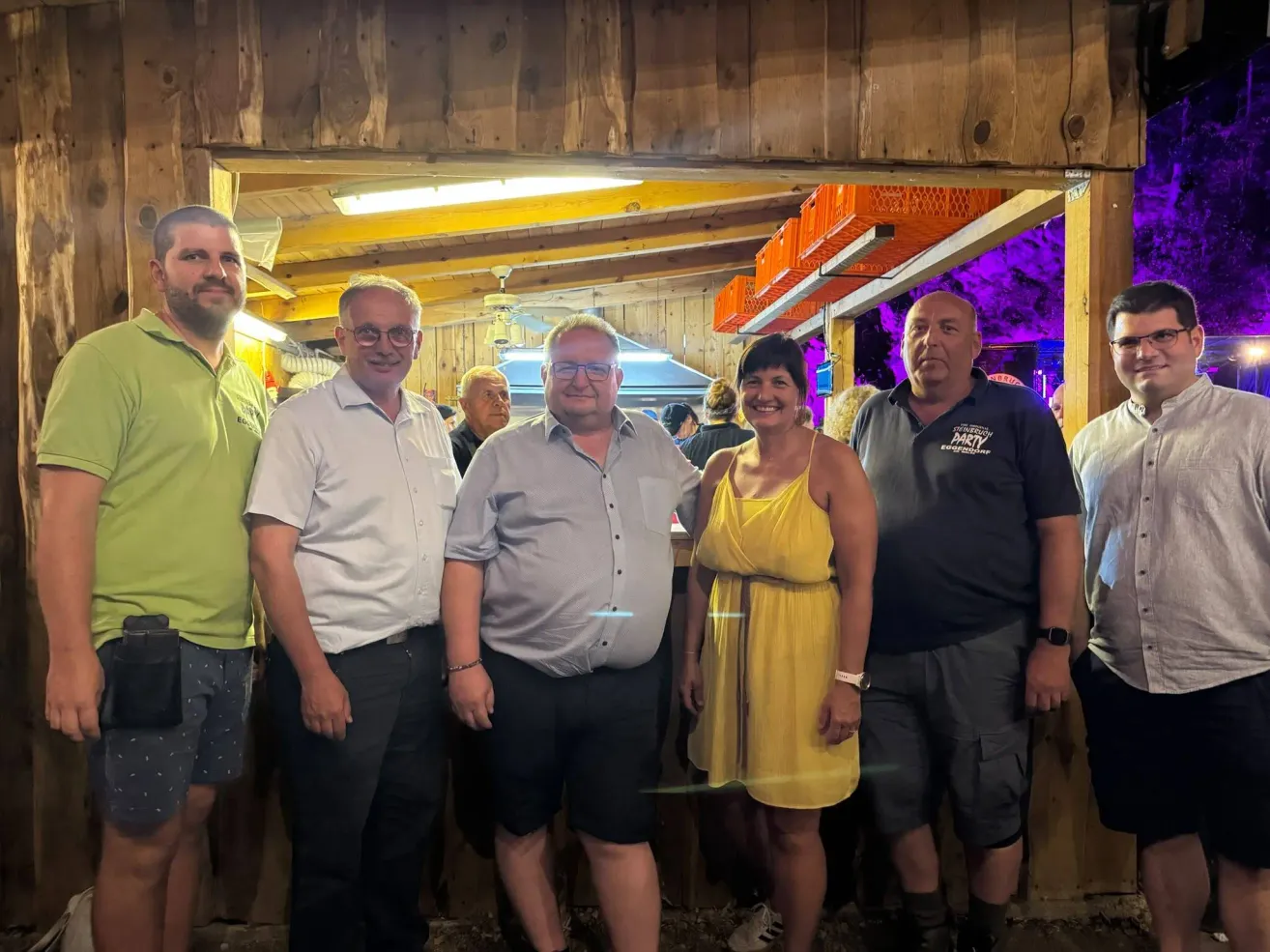 Fünf Erwachsene stehen in einer Gruppe in einer hölzernen Bar und lächeln für ein Foto. Der Mann in der Mitte trägt eine Brille und ein helles Hemd. Die Frau neben ihm trägt ein gelbes Kleid. Hinter ihnen ist eine hölzerne Bartheke zu sehen.