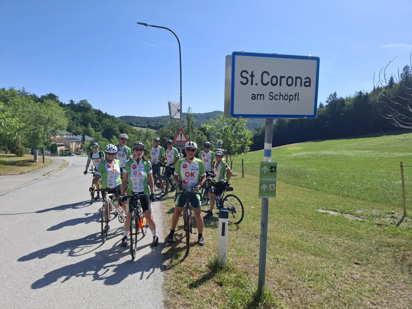 Eine Gruppe von Radfahrern in grünen und weißen Trikots mit 'OK' darauf steht für ein Foto in der Nähe eines Schildes, das 'St. Corona am Schöpfl' besagt.