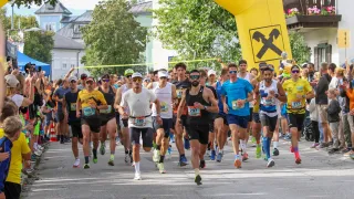 Eine Gruppe von Läufern nimmt an einem Marathon teil und läuft unter einem gelben Bogen mit dem Wort 'ffeisen' und einem Kreuzsymbol. Sie sind von Zuschauern und Bäumen umgeben.