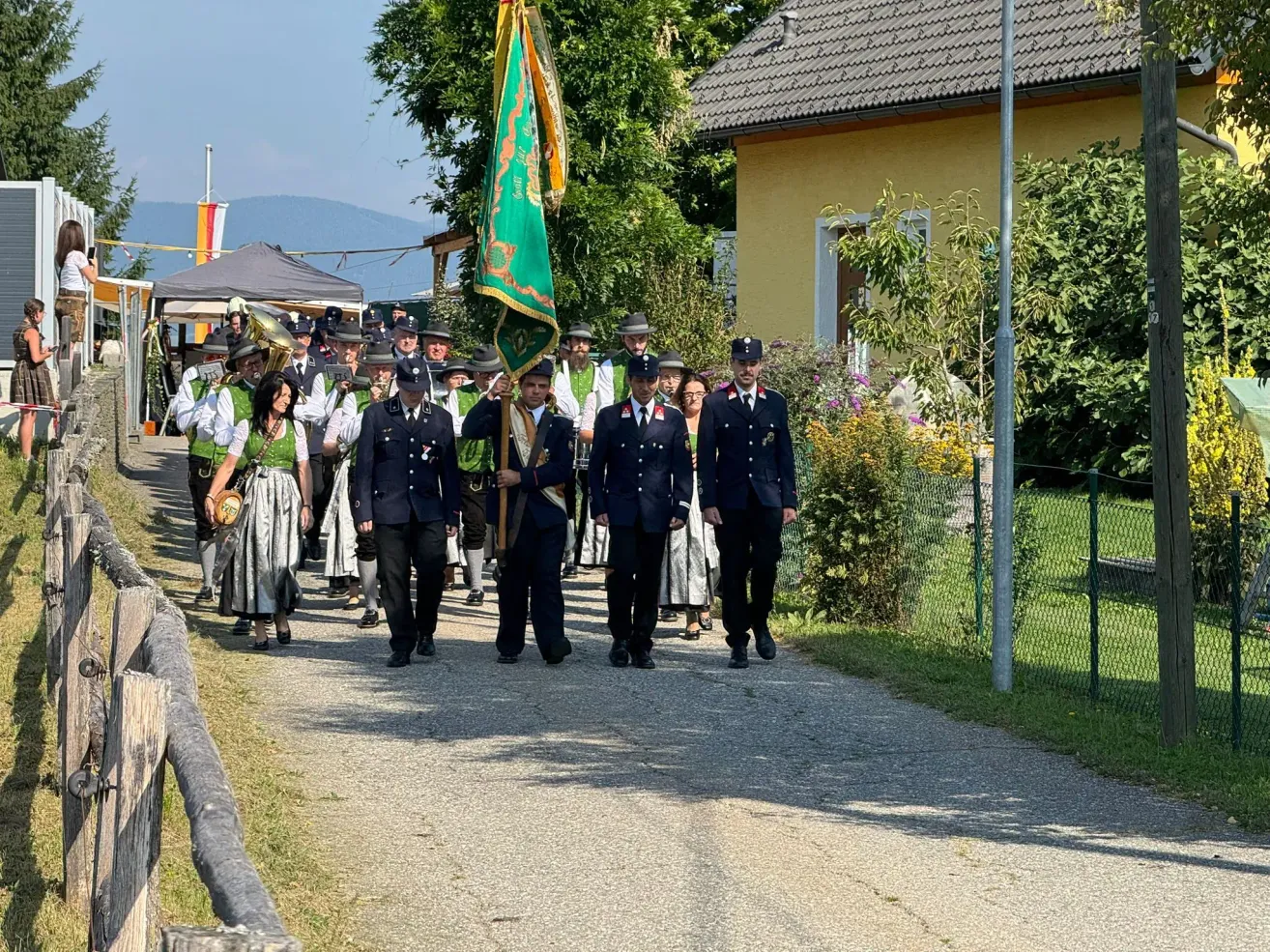 A group of people in uniform, including police officers, marching in a parade with a flag and a brass band, in front of a yellow house.