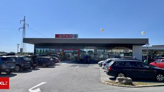 Ein SPAR-Supermarkt mit klarem blauem Himmel und einigen Autos davor. Eine Person geht in der Nähe des Eingangs.